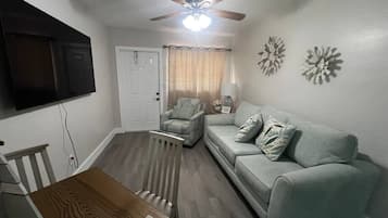 A living room with a ceiling fan, a television mounted on the wall, a sofa, a chair, and a dining table.