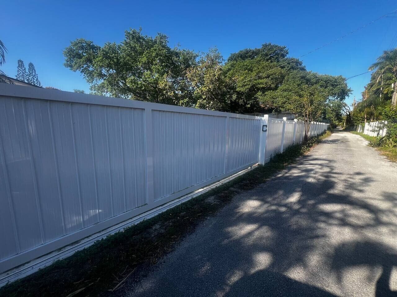 Una cerca blanca al borde de un camino pavimentado, con árboles al fondo.