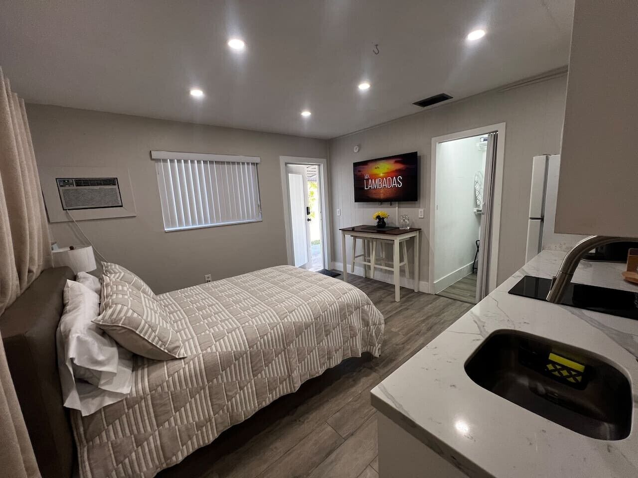 Un dormitorio con cama, televisor, una mesita y un área de cocina con lavaplatos.