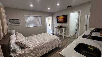 A bedroom with a bed, a TV, a small table, and a kitchen area with a sink.