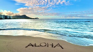Ein Strand mit 'ALOHA' im Sand geschrieben, Wellen und einem Berg im Hintergrund.