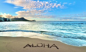 Ein Strand mit 'ALOHA' im Sand geschrieben, Wellen und einem Berg im Hintergrund.