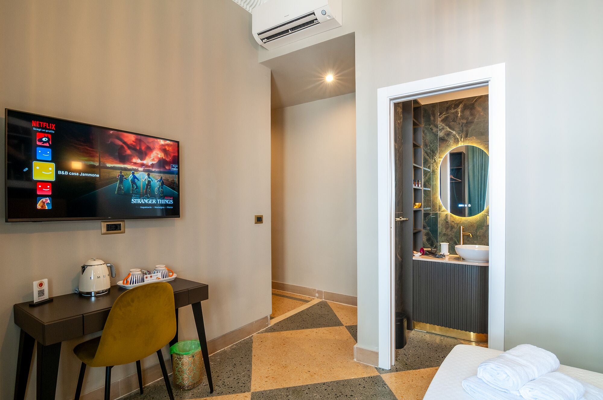 A hotel room with a TV displaying a movie, a desk with a coffee maker, a chair, and a bed with a neatly made white towel.