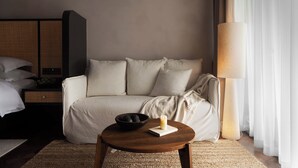A neutral-colored room with a bed, a sofa, a round wooden table with a candle and a bowl of fruit, and a lamp.