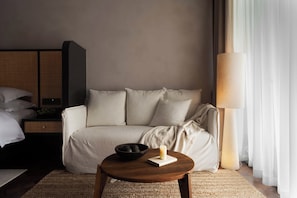 A neutral-colored room with a bed, a sofa, a round wooden table with a candle and a bowl of fruit, and a lamp.