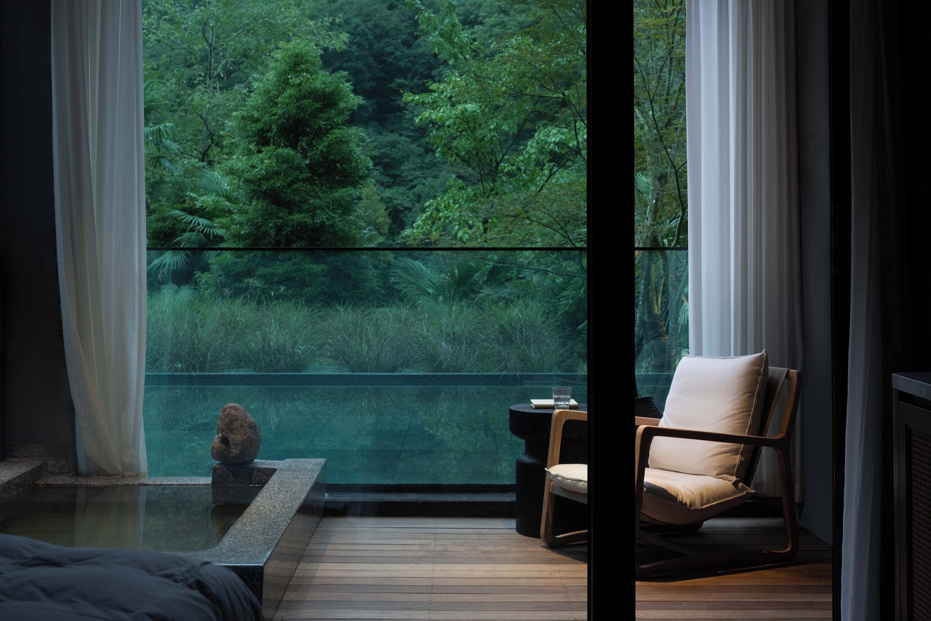 A room with a large window overlooking a forested area, a wooden floor, a chair, and a small table with a glass.