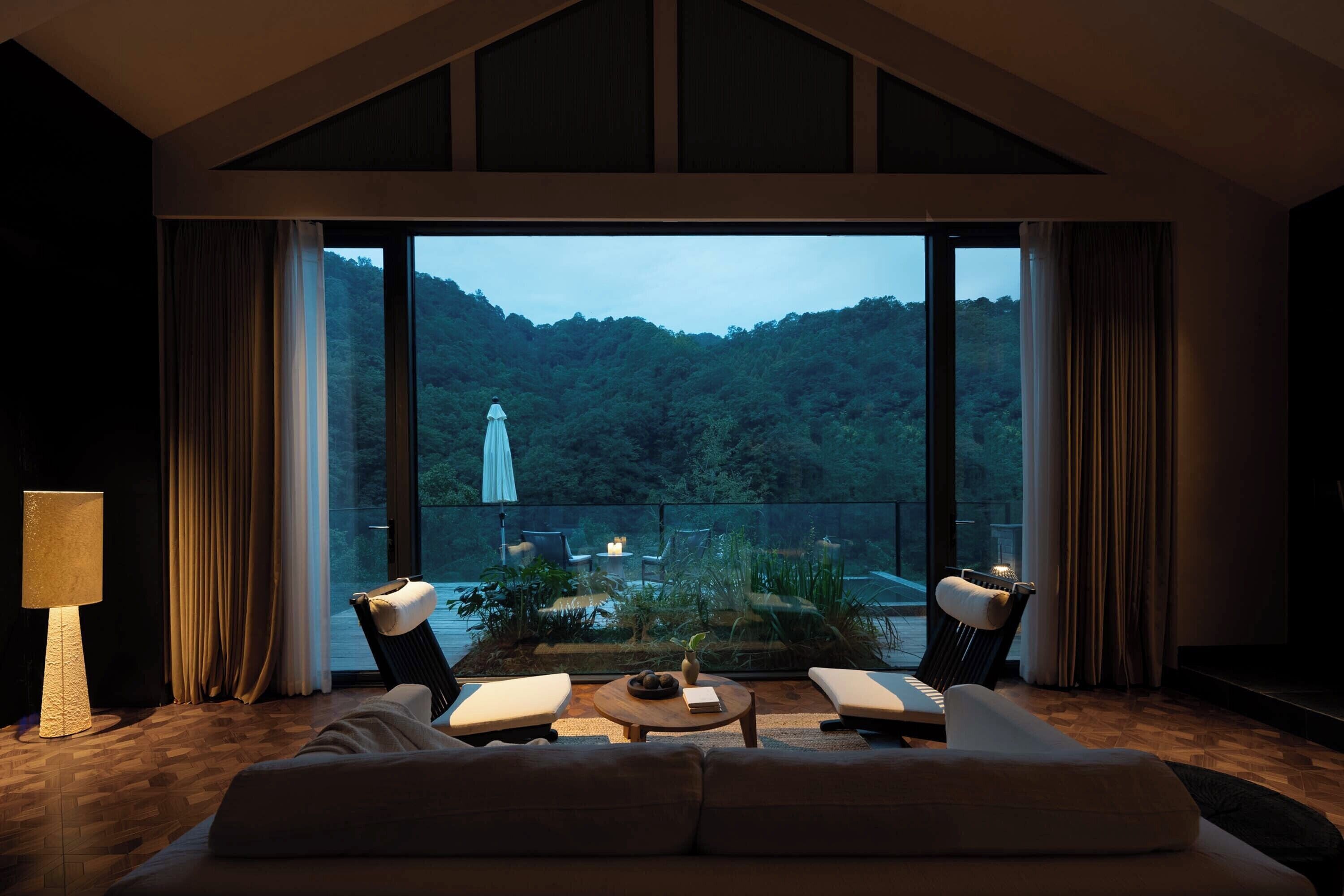 A spacious living room with a large window, a sofa, two armchairs, a coffee table, and a lamp.