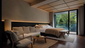 A modern hotel room with a large bed, a sofa, a coffee table, and a view of a lush garden.