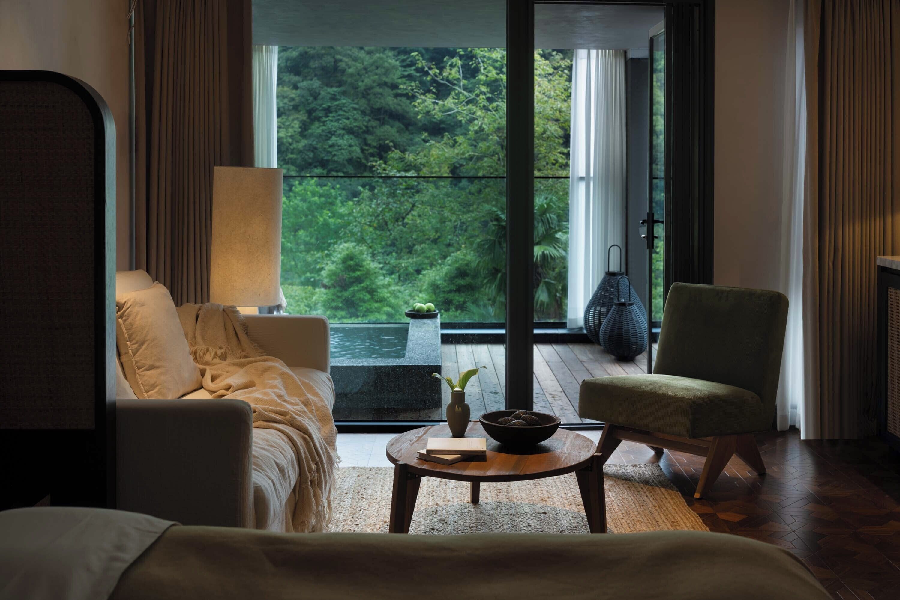 A room with a bed, a sofa, a chair, a small table, and a view of a lush green landscape through large windows.