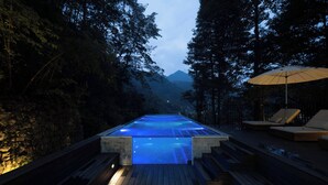 An infinity pool with a view of a mountain range, surrounded by trees and a stone wall.