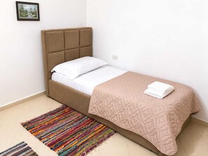 Ein Einzelbett mit Kopfteil, weißer Bettwäsche, einer beigen gesteppten Decke und einem farbenfrohen Teppich.