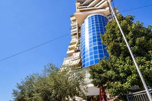 Ein hohes Gebäude mit einem blauen, zylindrischen Abschnitt, umgeben von Bäumen und einem wolkenlosen Himmel.