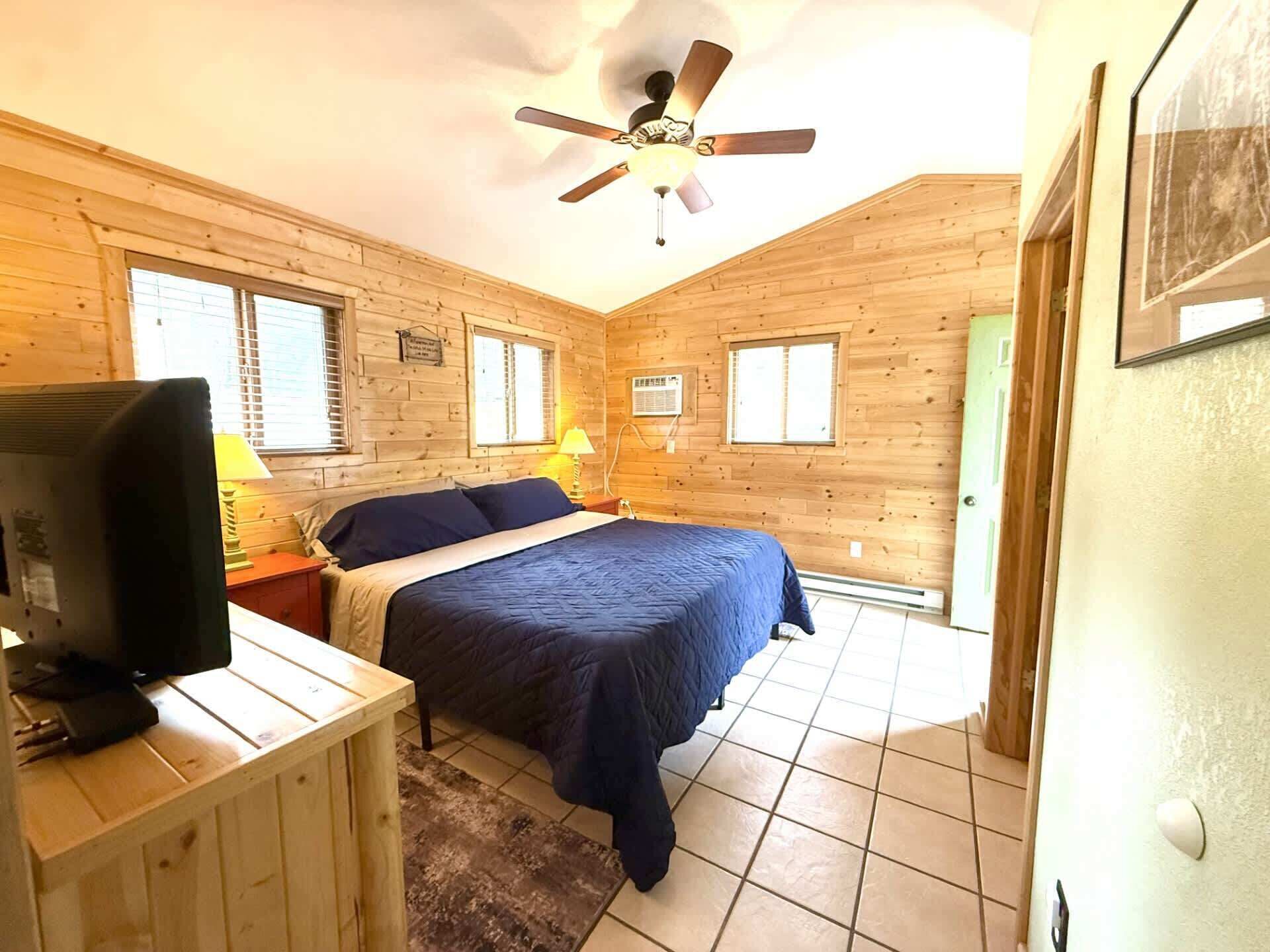 Ein Schlafzimmer in einer Holzhütte mit einem Bett, einem Schreibtisch, einem Fernseher und einem Deckenventilator.