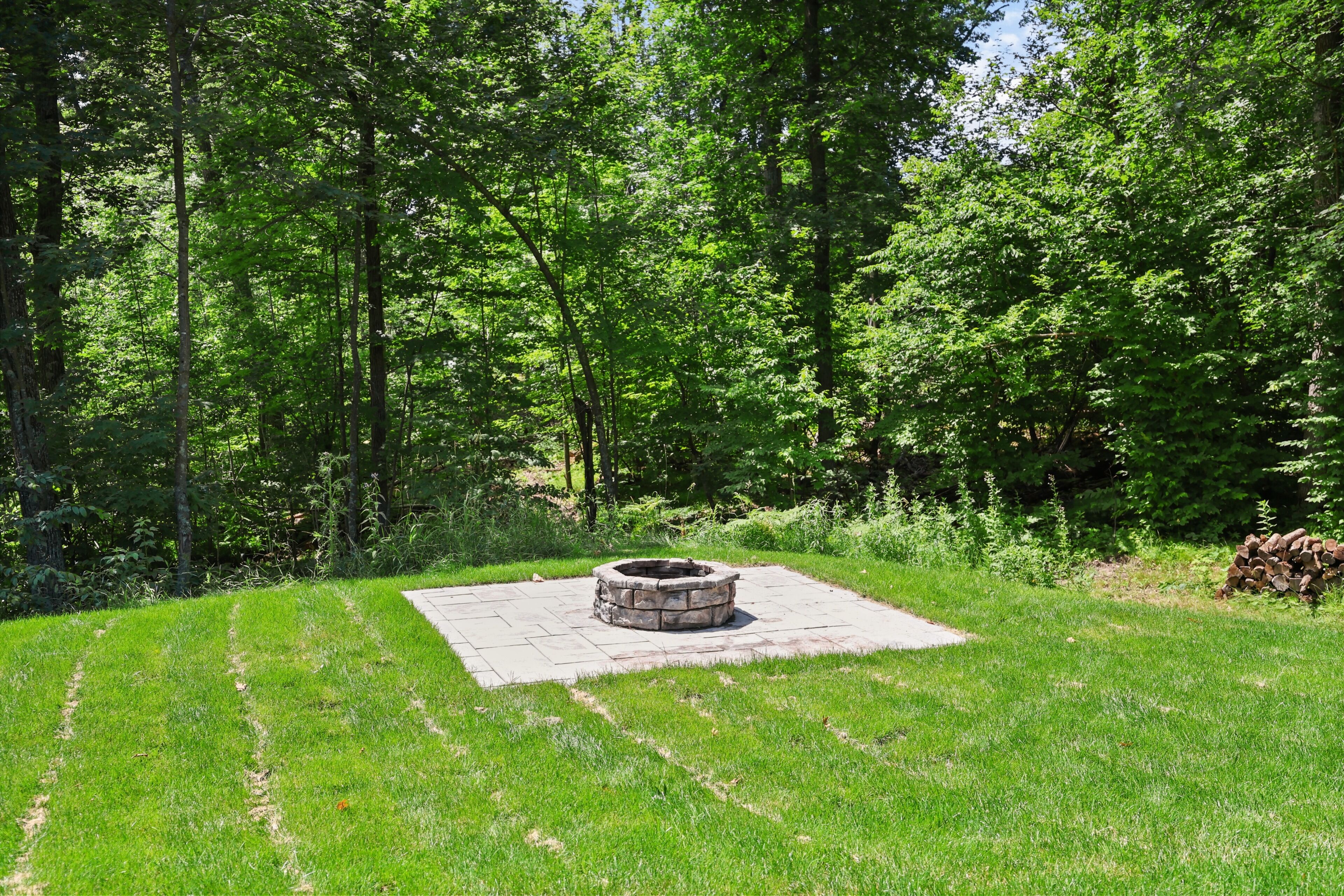 Un foyer en pierre, entouré d’une zone pavée, avec une pelouse verdoyante et des arbres touffus en arrière-plan.