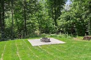 A stone fire pit surrounded by a paved area, with a grassy lawn and dense trees in the background. - Luxury Woodland Retreat Near Gull Lake, championship golf & Downtown Nisswa (Nisswa)