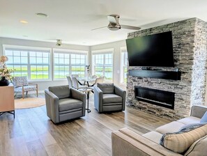 A spacious living room with a stone fireplace, a flat-screen TV, leather sofas, and large windows offering a view of the outdoors. - 22 South Shore Drive Ext - South Shore Marina (Bethany Beach)
