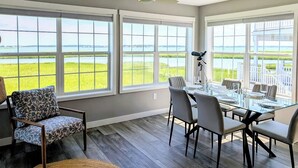A dining area with a glass table and chairs, large windows with a view of a grassy landscape, and a ceiling fan.