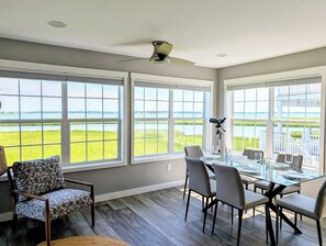 A dining area with a glass table and chairs, large windows with a view of a grassy landscape, and a ceiling fan. - 22 South Shore Drive Ext - South Shore Marina (Bethany Beach)