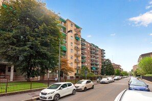 Un immeuble résidentiel à plusieurs étages, doté de balcons, avec des voitures garées le long de la rue et des arbres bordant le trottoir.