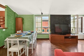 A dining area with a table set for four, a television on a wooden stand, and a view of a building outside through a window. - Spacious and bright penthouse, ideal for up to 7 people (Genova)
