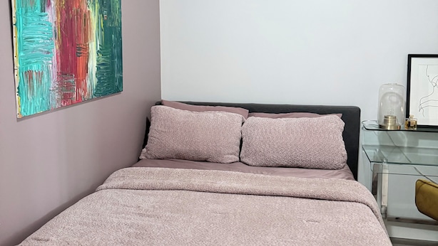 Un dormitorio con una cama, una mesita de vidrio, una silla y un cuadro abstracto colorido en la pared.