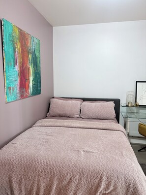 A bedroom with a bed, a glass side table, a chair, and a colorful abstract painting on the wall.