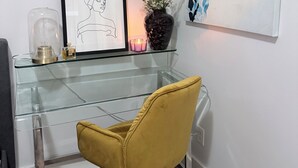 Una habitación moderna con un escritorio de vidrio, una obra de arte enmarco, un jarrón con flores y una silla amarilla.