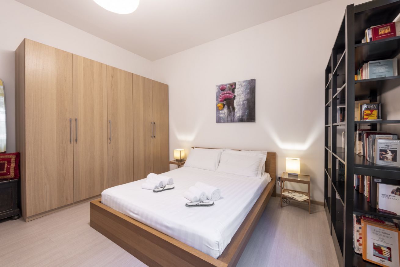 A bedroom with a bed, a wardrobe, a bedside table with a lamp, and a bookshelf.