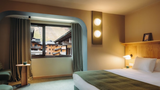 Habitación de hotel con cama, mesita de noche, lámpara, ventana con cortinas y vistas a edificios.