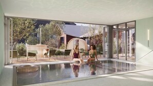 Piscina interior moderna con paredes de vidrio, una persona sentada junto a la piscina y mobiliario exterior.