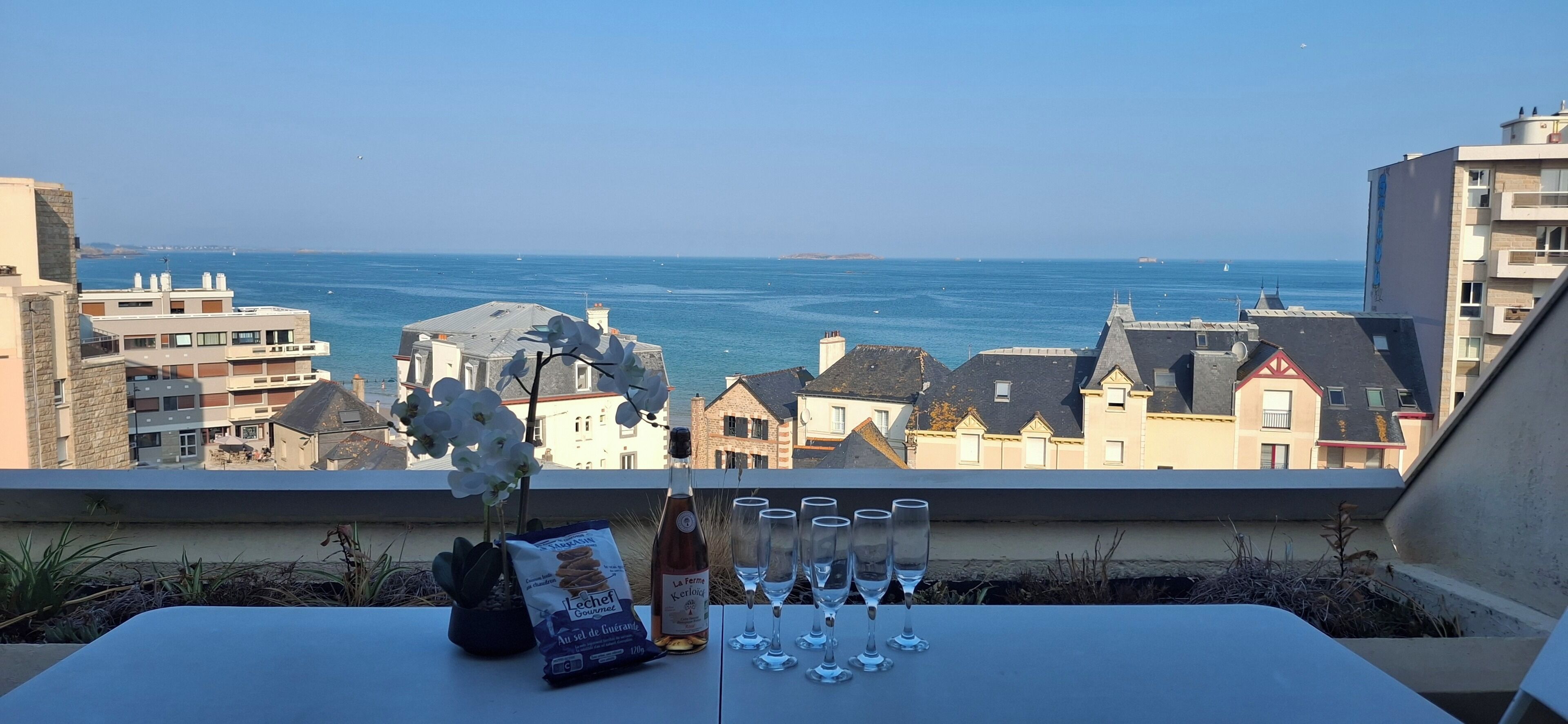 A table with a bottle, glasses, and a bag of crackers, overlooking a coastal view with buildings and the sea.