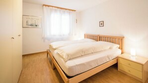 Un dormitorio con cama, mesita de noche, cómoda, ventana con cortinas e imagen colgada en la pared.