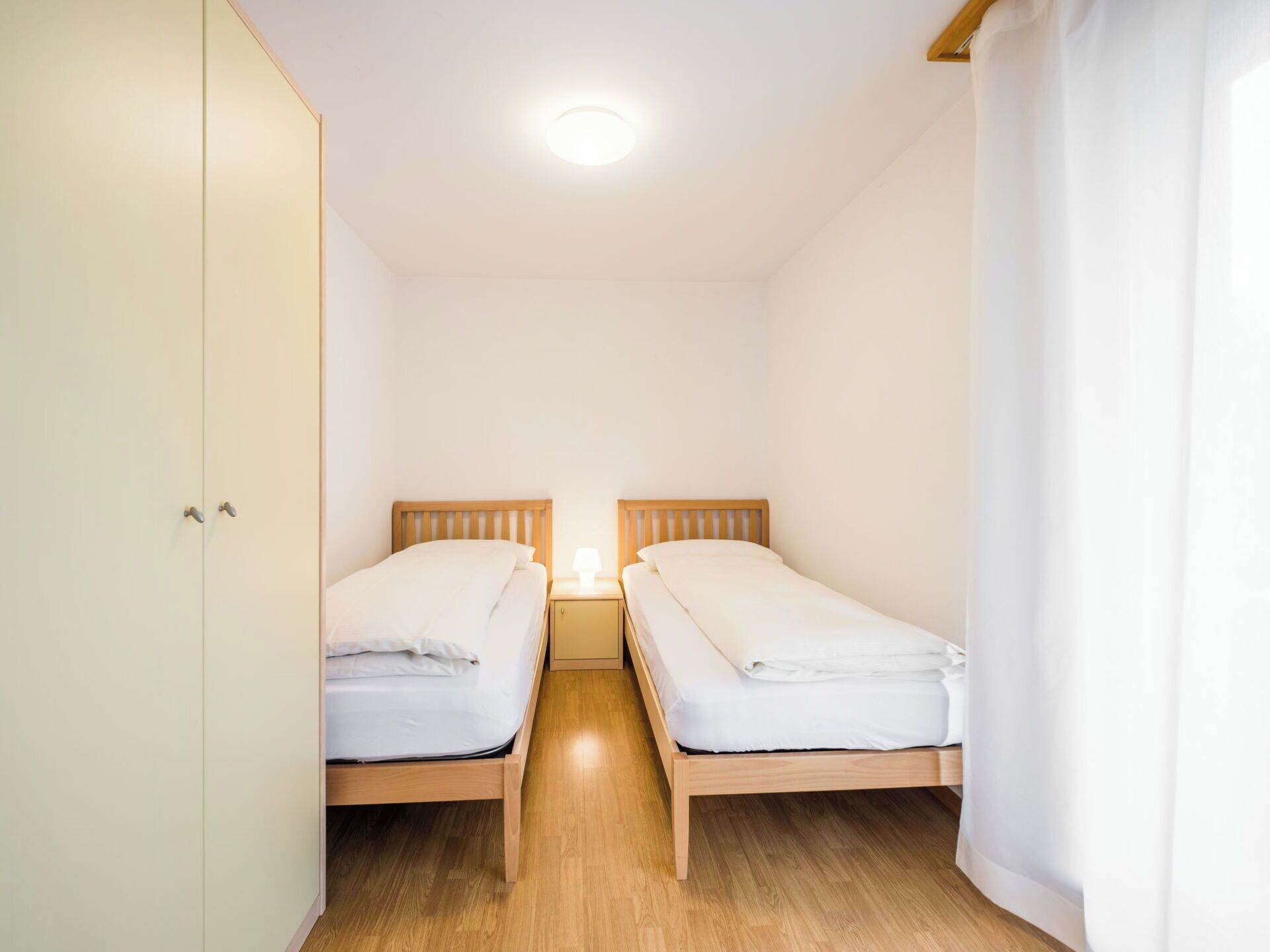 Een kleine, schone kamer met twee eenpersoonsbedden, een kledingkast en een nachtkastje met een lamp.