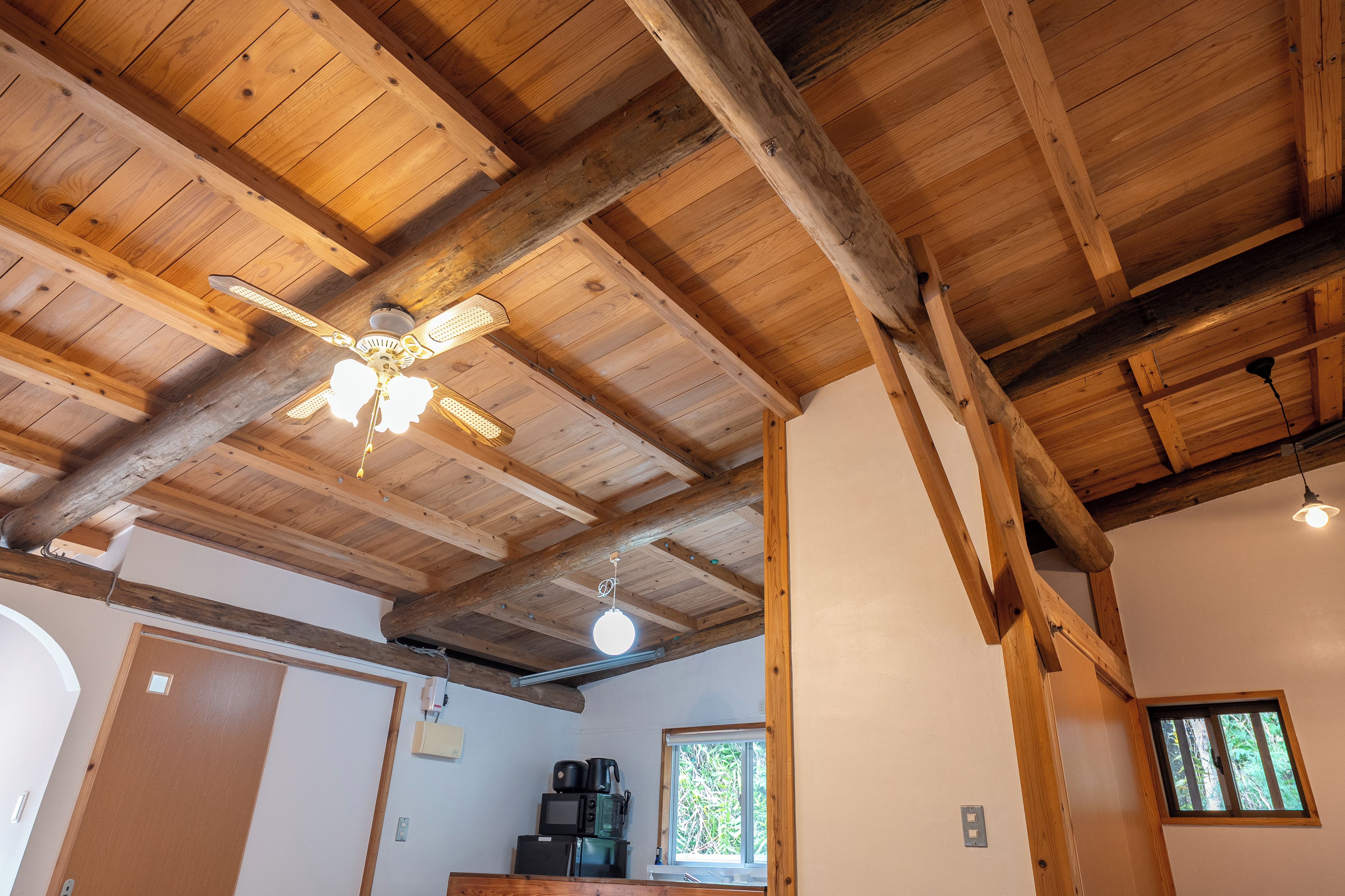 A room with wooden beams, a ceiling fan, a window, and a kitchenette with appliances.