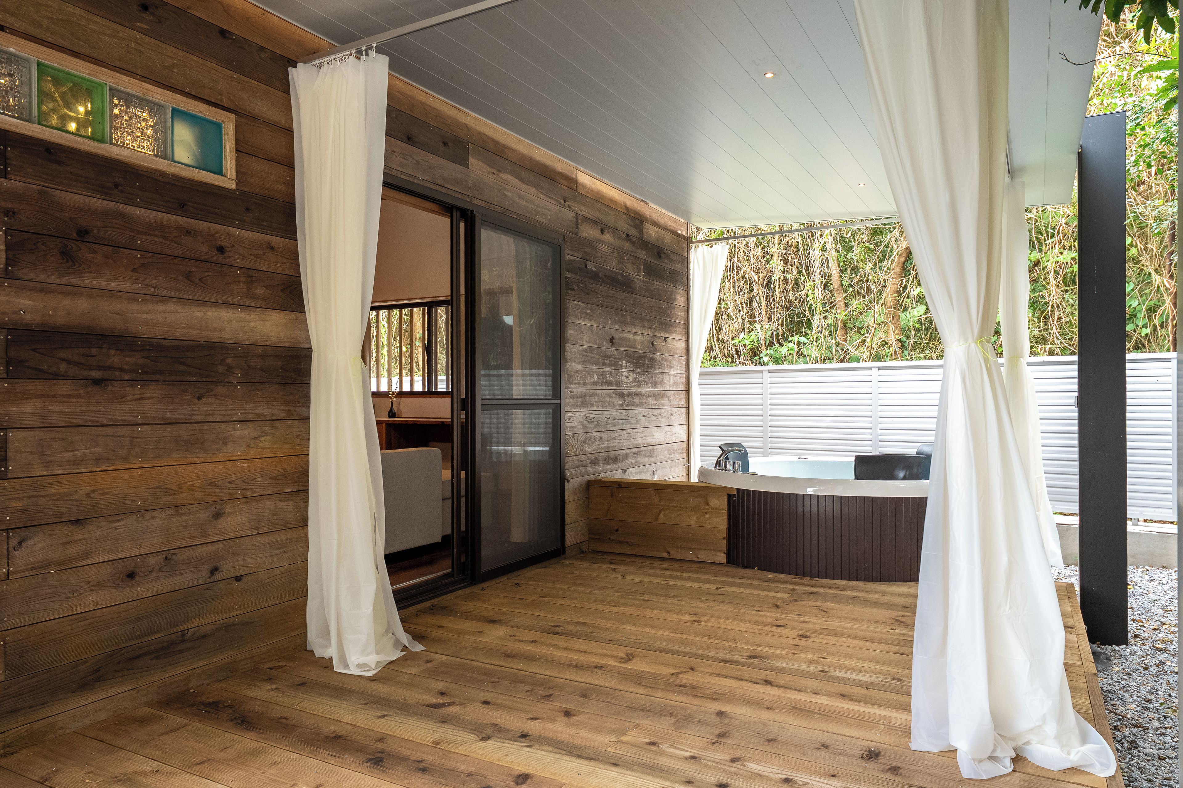 Una terraza de madera con un ventanal que da a una habitación, un jacuzzi y una zona cubierta con cortinas.