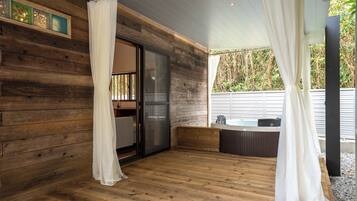 Una terraza de madera con un ventanal que da a una habitación, un jacuzzi y una zona cubierta con cortinas.