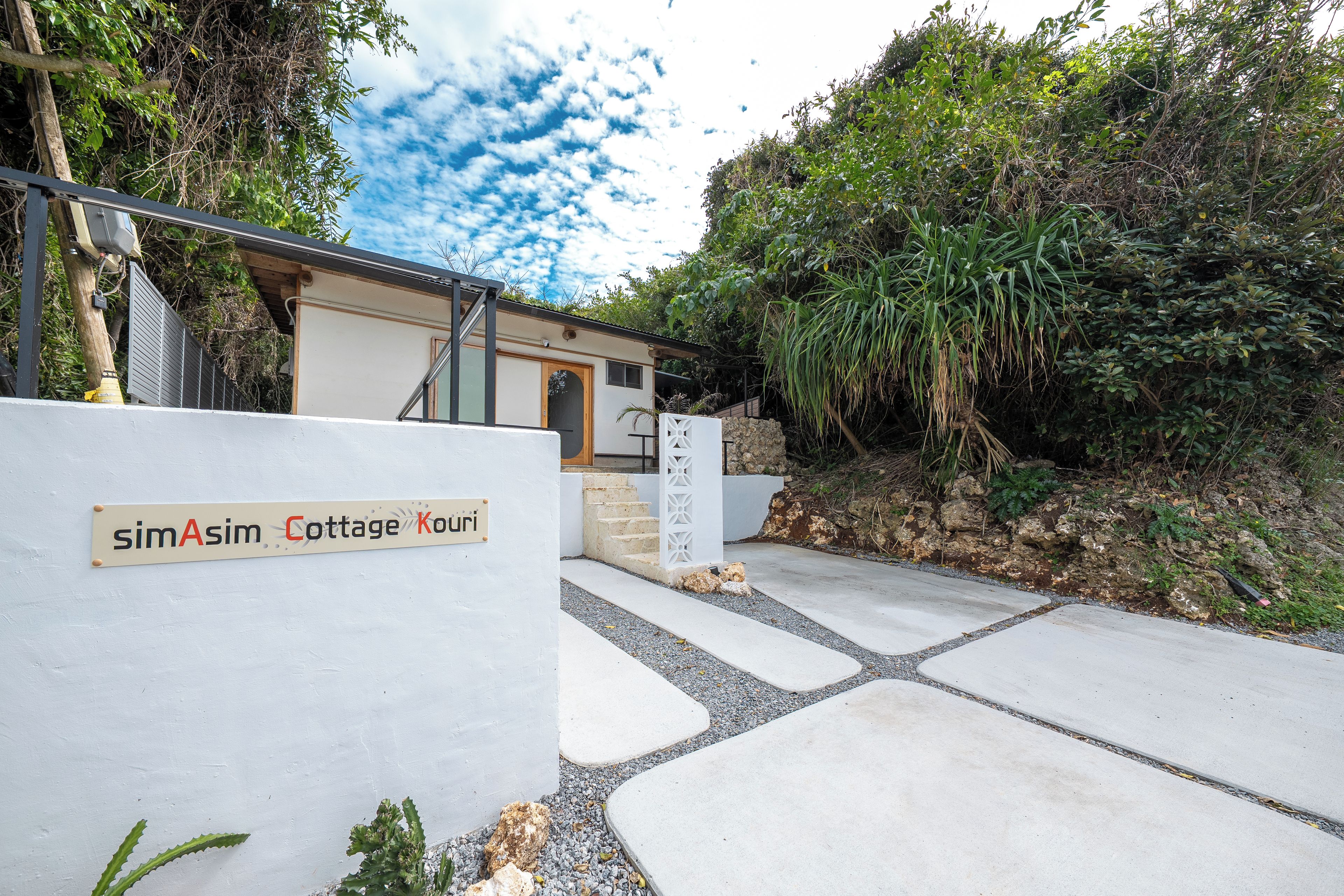 Entrada a una cabaña con un letrero que dice 'simAsim Cottage Kouri', un camino de hormigón y follaje circundante.