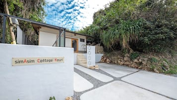 A cottage entrance with a sign reading 'simAsim Cottage Kouri,' a concrete pathway, and surrounding greenery.
