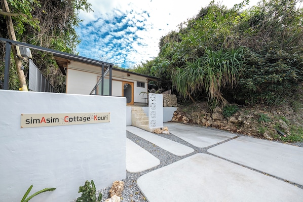 Entrada a una cabaña con un letrero que dice 'simAsim Cottage Kouri', un camino de hormigón y follaje circundante.