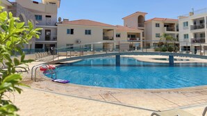 A swimming pool with a surrounding deck, clear skies, and multi-story buildings in the background. - Family Resort Apt with Pool Sleeps 6 (Paralimni)