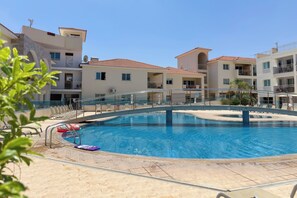 A swimming pool with a surrounding deck, clear skies, and multi-story buildings in the background. - Family Resort Apt with Pool Sleeps 6 (Paralimni)