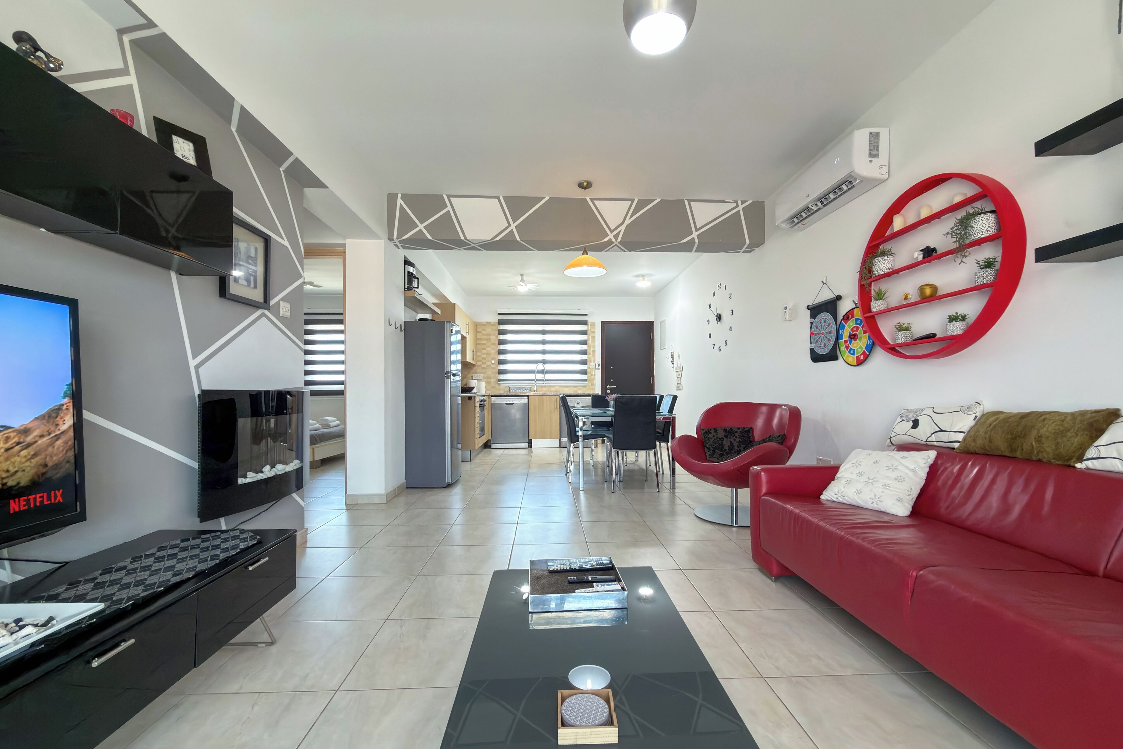 A modern living room with a flat-screen TV, a red sofa, a coffee table, and a dining area with a kitchen in the background.