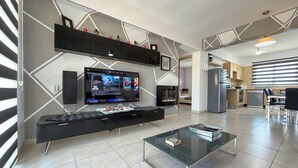 A modern living room with a flat-screen TV, a glass coffee table, and a kitchen area in the background. - Family Resort Apt with Pool Sleeps 6 (Paralimni)