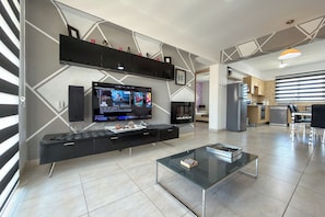 A modern living room with a flat-screen TV, a glass coffee table, and a kitchen area in the background. - Family Resort Apt with Pool Sleeps 6 (Paralimni)