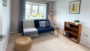 Een woonkamer met een bank, een klein rond voetenbankje, een boekenkast en een raam met gordijnen.