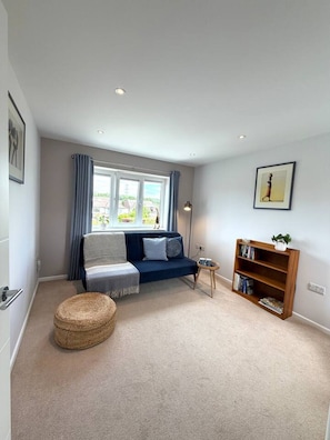 A living room with a sofa, a small round ottoman, a bookshelf, and a window with curtains. - Modern Annexe, 10 Minutes from Oxford City Centre (Oxford)