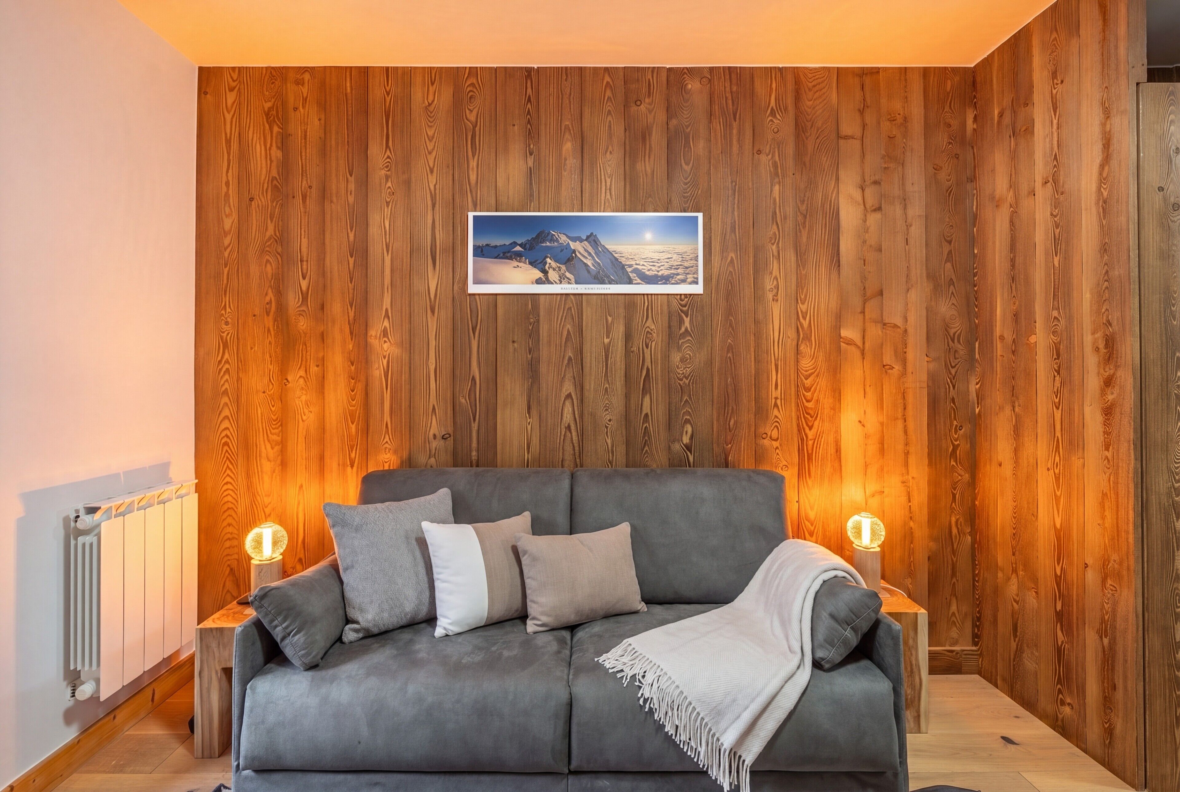 A cozy room with a grey sofa, wooden panel walls, a framed picture, and two bedside lamps.