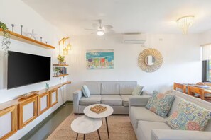 Ein modernes Wohnzimmer mit Flachbildfernseher, einem grauen Sofa, einem Couchtisch und einem Essbereich mit Holztisch und Stühlen.