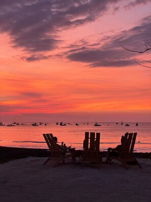 Ein Strandbild mit drei Holzstühlen, die zum Meer hin zeigen, bei Sonnenuntergang.