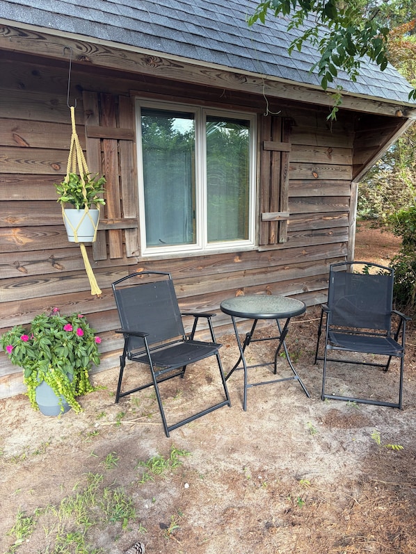 Une cabane en bois avec une fenêtre, une plante suspendue et un petit espace extérieur aménagé avec deux chaises et une table.